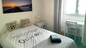 A neatly arranged bedroom with a bed, pillows, a nightstand, a lamp, a chair, and a window with curtains. - Renovated house in town center, 3 bedrooms, near shops (Groix)