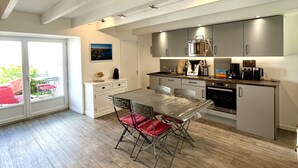 A modern kitchen with stainless steel appliances, a dining area with a glass table, and a view of the outdoors through a sliding door. - Renovated house in town center, 3 bedrooms, near shops (Groix)