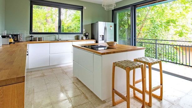 A modern kitchen with a large island, white cabinetry, and a view of lush greenery through large windows.
