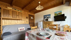 Ein gemütliches Zimmer mit Holzboden, einer Couch mit Kissen, einem Tisch gedeckt mit Tellern und Gläsern sowie einem an der Wand befestigten Fernseher.