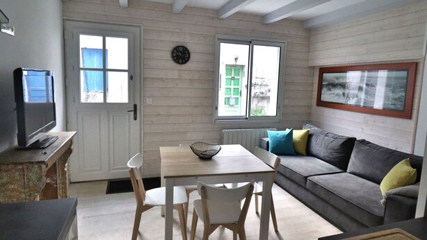Een woonkamer met een bank, een tafel en stoelen. Er is een televisie op een meubel, een klok aan de muur en een raam met uitzicht op een gebouw.