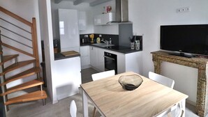 Een moderne keuken met witte kastjes, een houten trap, een eettafel met een zwarte kom en een televisie op een stenen console.