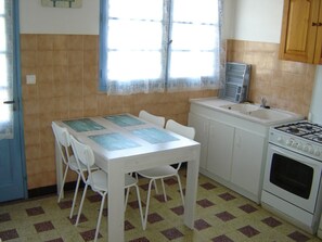 Una cocina pequeña con una mesa blanca, cuatro sillas, una estufa, un lavabo y una ventana con cortinas.