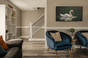 Un salon avec un escalier, un canapé, deux fauteuils, une bibliothèque et un tableau représentant un cygne.