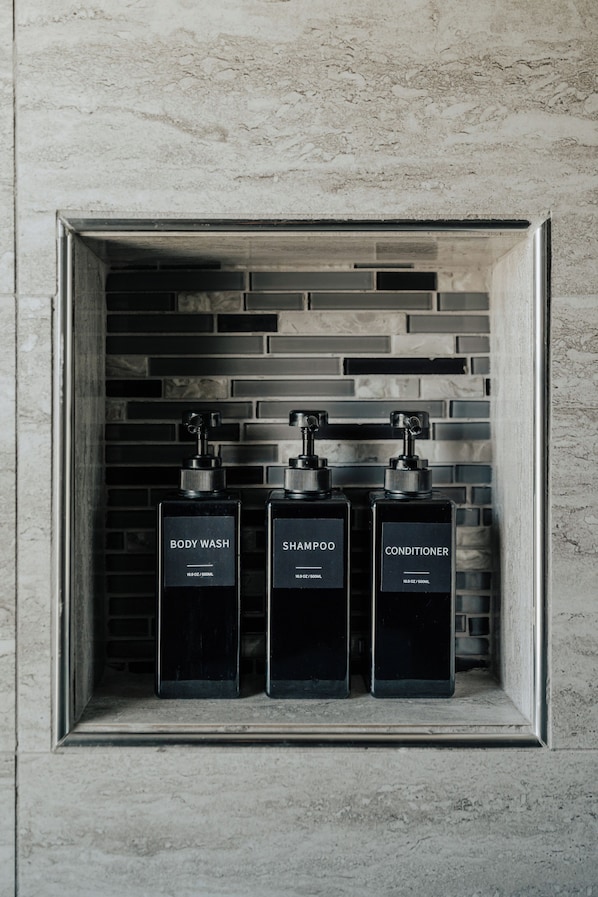 Un baño con pared de azulejos y un estante que contiene tres botellas negras etiquetadas como “BODY WASH”, “SHAMPOO” y “CONDITIONER”.
