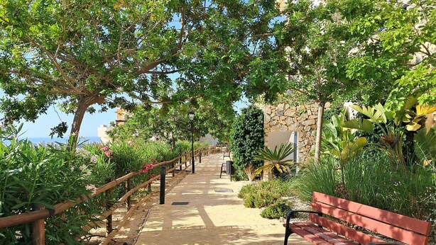 Un camí pavimentat delimitat per baranes de fusta i amb bancs, que condueix cap a un edifici amb façana de pedra.