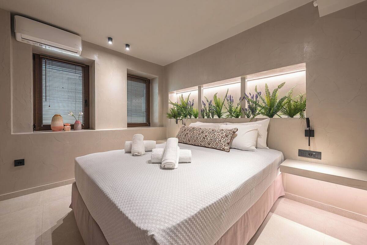 A modern bedroom with a large bed, a built-in window with blinds, a wall-mounted air conditioner, and a bedside plant display.