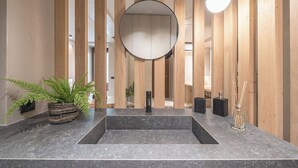 Ein modernes Badezimmer mit einem rechteckigen Waschbecken, einem runden Spiegel, Holzlamellen, einem Topf mit Pflanze und einer Pendelleuchte.