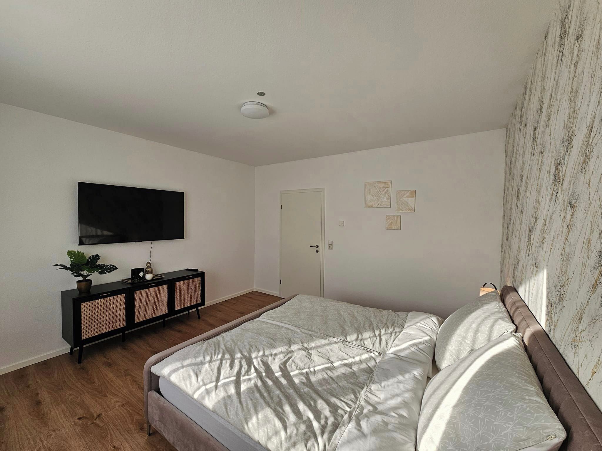 Un dormitorio con una cama, un televisor montado en la pared, una puerta y un mueble bajo con una planta.