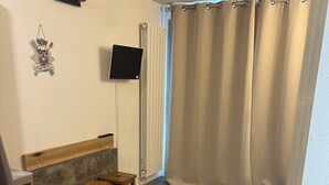 Une pièce dotée d’un plancher en bois, d’un téléviseur fixé au mur, d’une fenêtre avec des rideaux et d’un petit coin salon avec des tabourets.