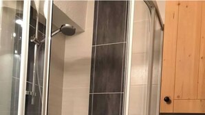 Une douche avec une porte en verre, un mur recouvert de carreaux noirs verticaux et un meuble en bois sur le côté droit.