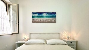 A bedroom with a bed, two bedside tables, a nightstand, a window, and a wall-mounted painting of a beach scene.