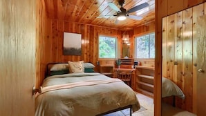 A wooden cabin bedroom with a bed, a desk, and a chair. There is a window and a door visible.