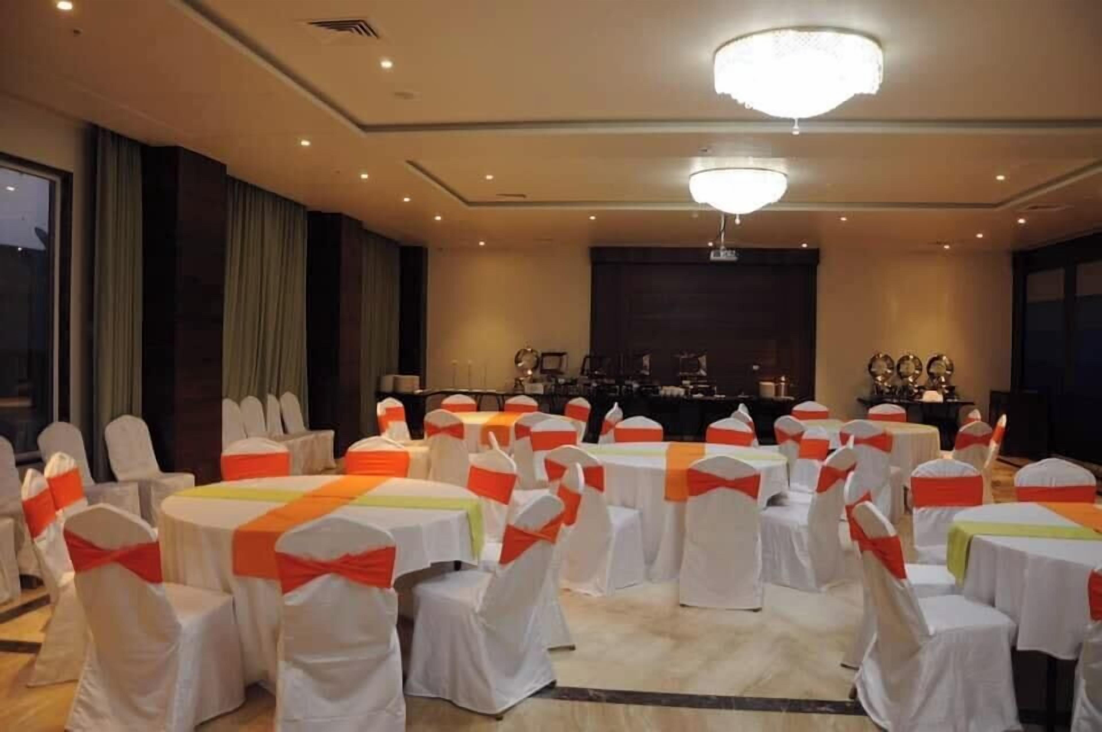 A conference room with round tables, white chairs with orange sashes, a stage with speakers, and a chandelier.