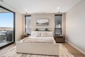 A modern bedroom with a large bed, bedside tables, a painting, and a view through a glass door.