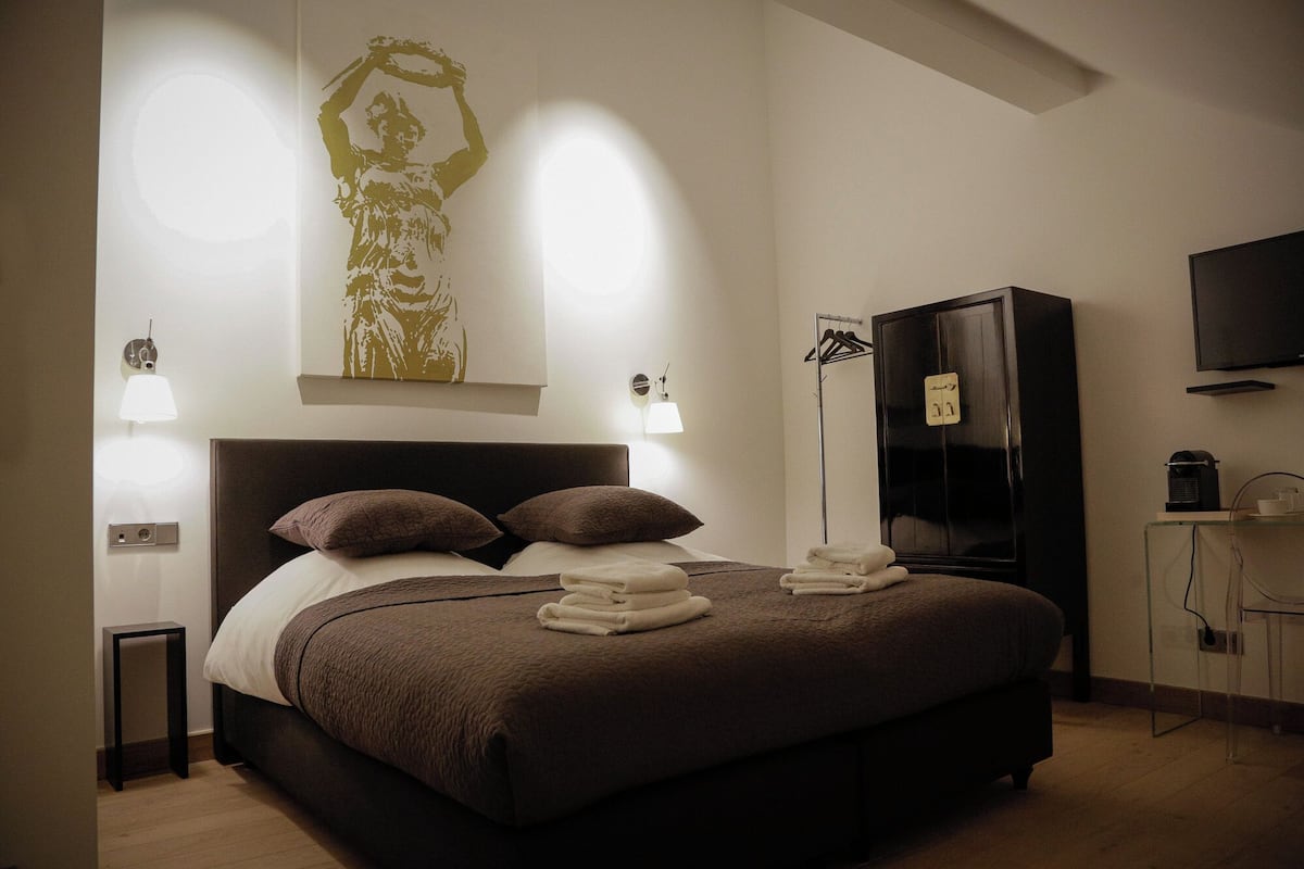 Un lit bien fait, agrémenté d’oreillers et de serviettes, une armoire noire, une lampe sur pied et un téléviseur fixé au mur.