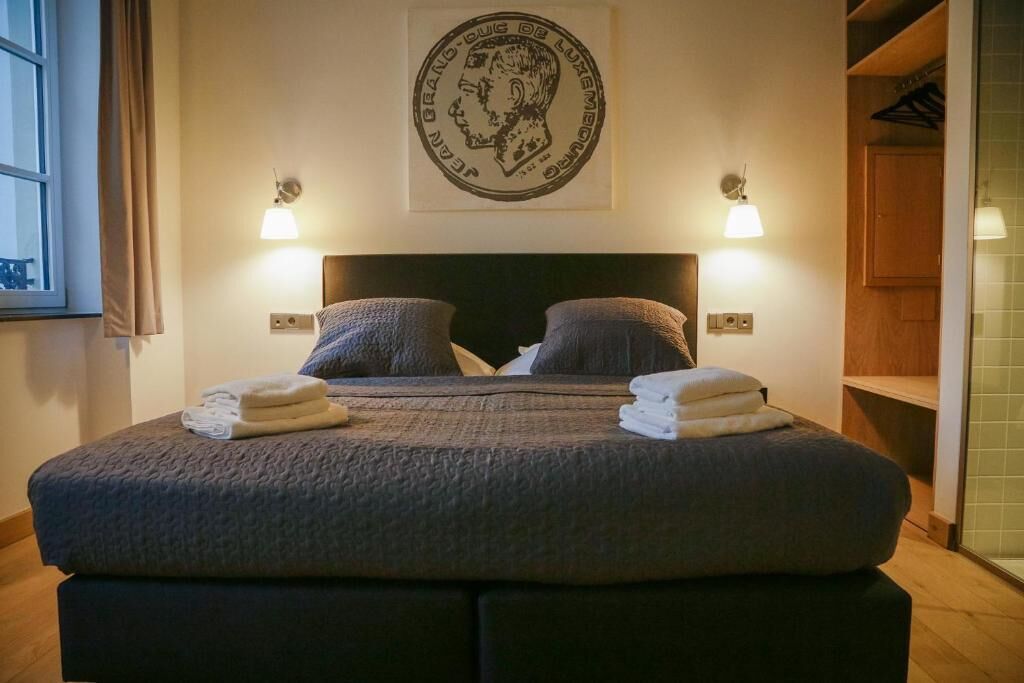 Een bed met een donker hoofdbord, twee kussens en twee opgevouwen handdoeken. Daarboven hangt een wandlamp en aan de muur een ingelijst kunstwerk.