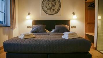 Een bed met een donker hoofdbord, twee kussens en twee opgevouwen handdoeken. Daarboven hangt een wandlamp en aan de muur een ingelijst kunstwerk.