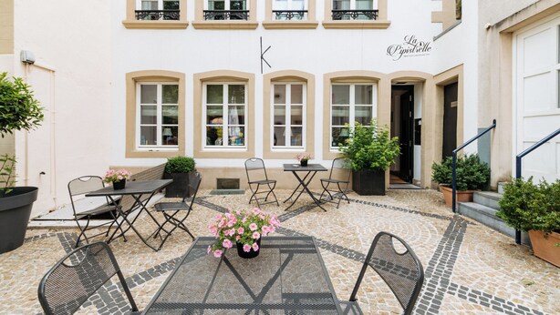Une cour avec des tables et des chaises à l’extérieur, des plantes en pot et un bâtiment doté de fenêtres en arc de cercle et d’une enseigne indiquant « La Pizzeria ».