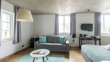 Une chambre d’hôtel moderne avec un canapé gris, une petite table ronde, un lit recouvert d’une couverture bleue, un bureau avec un ordinateur et un téléviseur fixé au mur.