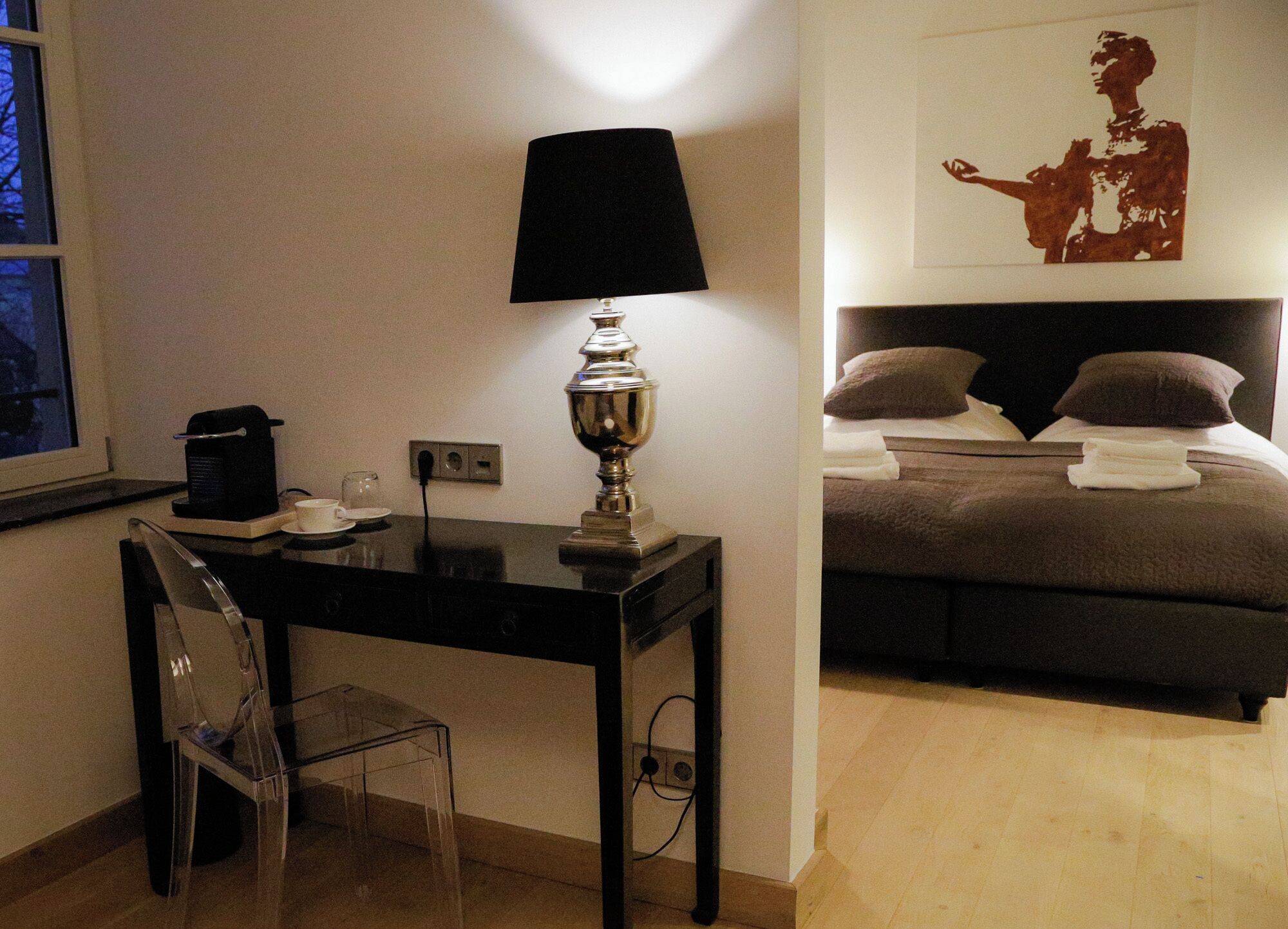 A hotel room with a bed, a bedside table with a lamp, a chair, and a coffee machine.