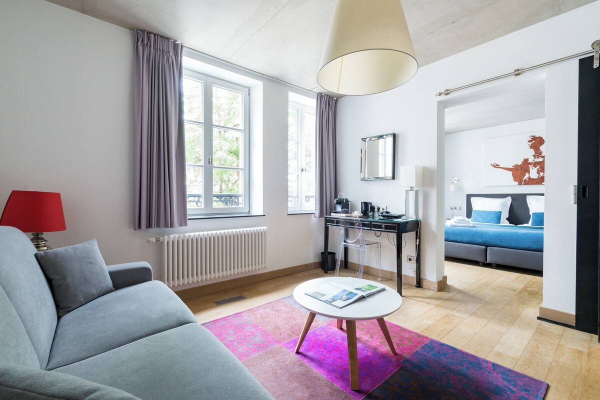 Une chambre d’hôtel moderne avec un canapé gris, une petite table ronde sur laquelle repose un magazine, un bureau avec une chaise, un lit avec une literie bleue et une grande fenêtre avec des rideaux.