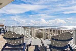 Una terraza con sillas Adirondack y una mesa con vista a una playa y una cerca blanca.