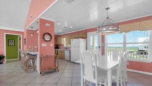 A spacious kitchen with white cabinets, a dining area with white chairs, and a view of the outdoors through large windows. - Beautiful Gulf Front Home With Amazing Views! (Gulf Shores)