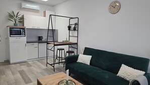 Un ambiente compatto con angolo cottura, un divano verde, un tavolino in legno e una sveglia a parete.