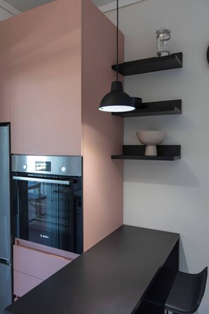 Una cocina con horno Bosch, armarios negros y rosas, una encimera negra y un lámpara colgante.