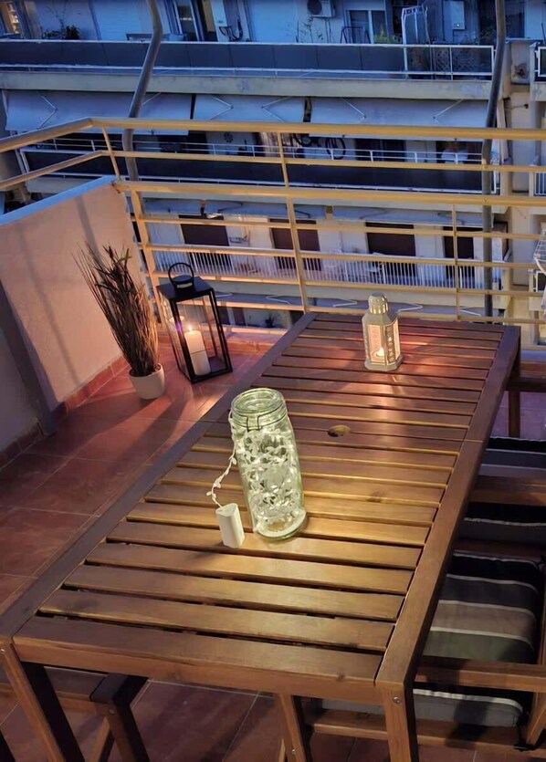 Una mesa de madera con un tarro de vidrio, dos velas y una planta en maceta en un balcón con barandillas y vistas a edificios.