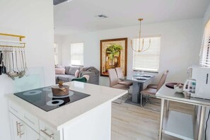 Cocina moderna con una isla central, comedor con mesa y sillas, un sofá y un ventanal con persianas.