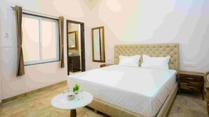 A bedroom with a large bed, a bedside table, a small round table with a plant, and a window with curtains. - Gulmohar Retreat And Spa (Kumbhalgarh)