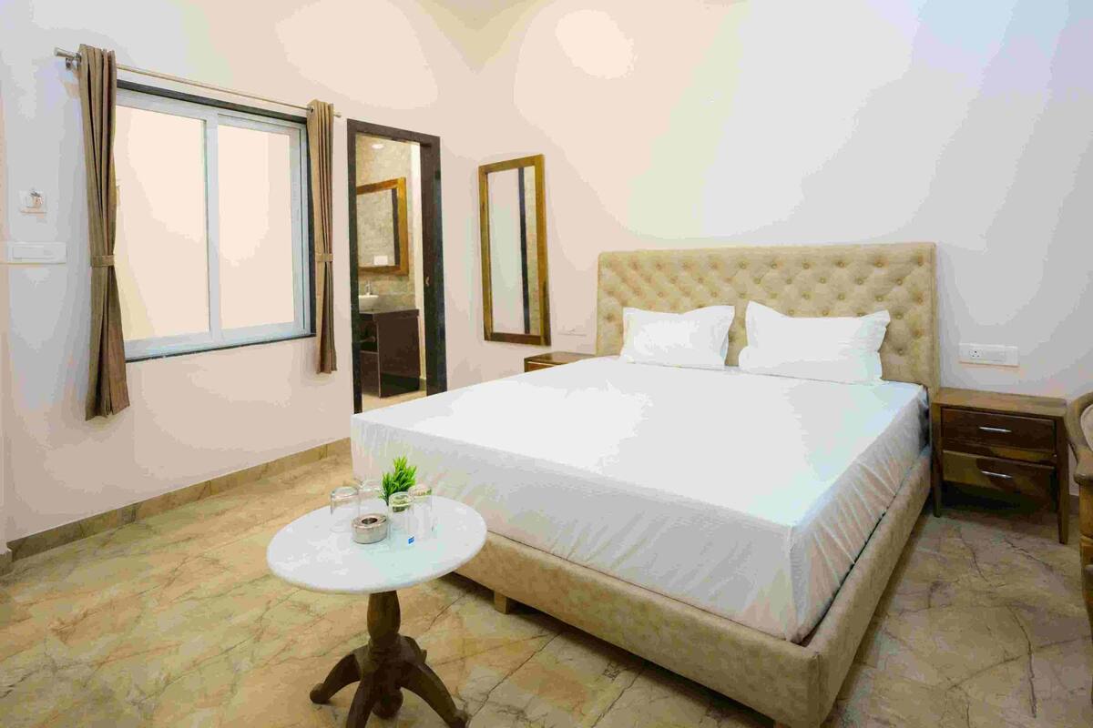 Un dormitorio con una cama grande, una mesita de noche, una pequeña mesa redonda con una planta y una ventana con cortinas.