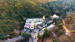 An aerial view of a hotel complex with multiple buildings, including a prominent dome structure, surrounded by lush greenery and a winding road. - Gulmohar Retreat And Spa (Kumbhalgarh)