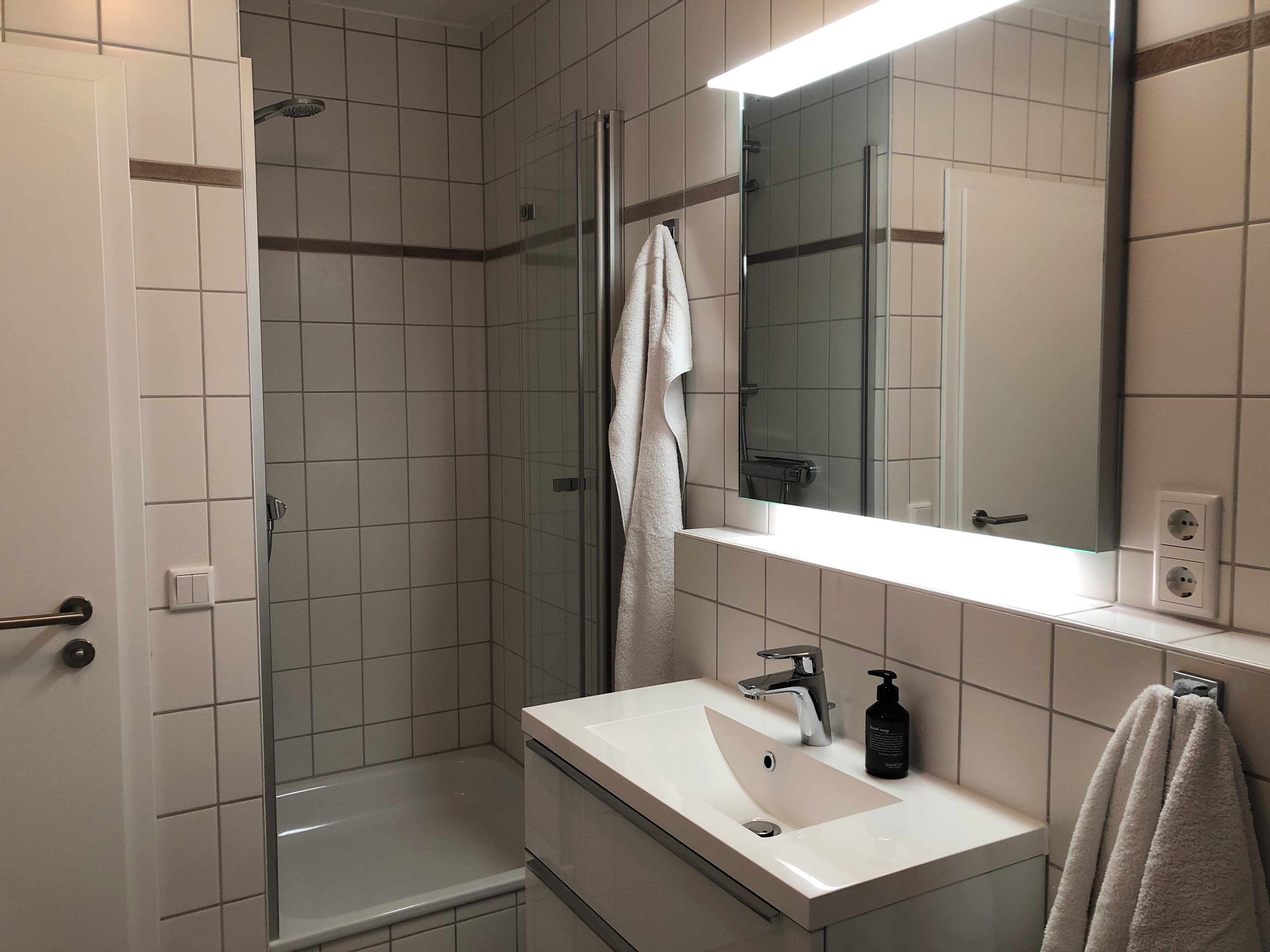 Ein modernes Badezimmer mit einer gläsernen Duschkabine, einem weißen Waschbecken mit Spiegel darüber und einem Handtuch an der Wand.