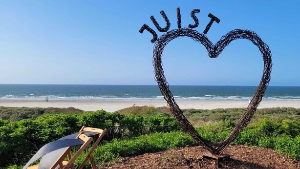 Scultura a forma di cuore realizzata con fili metallici intrecciati, con la scritta 'JUST' su uno sfondo di spiaggia.