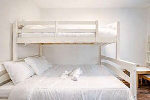 Una cama litera con ropa de cama y almohadas blancas, un banco de madera y una pared blanca.