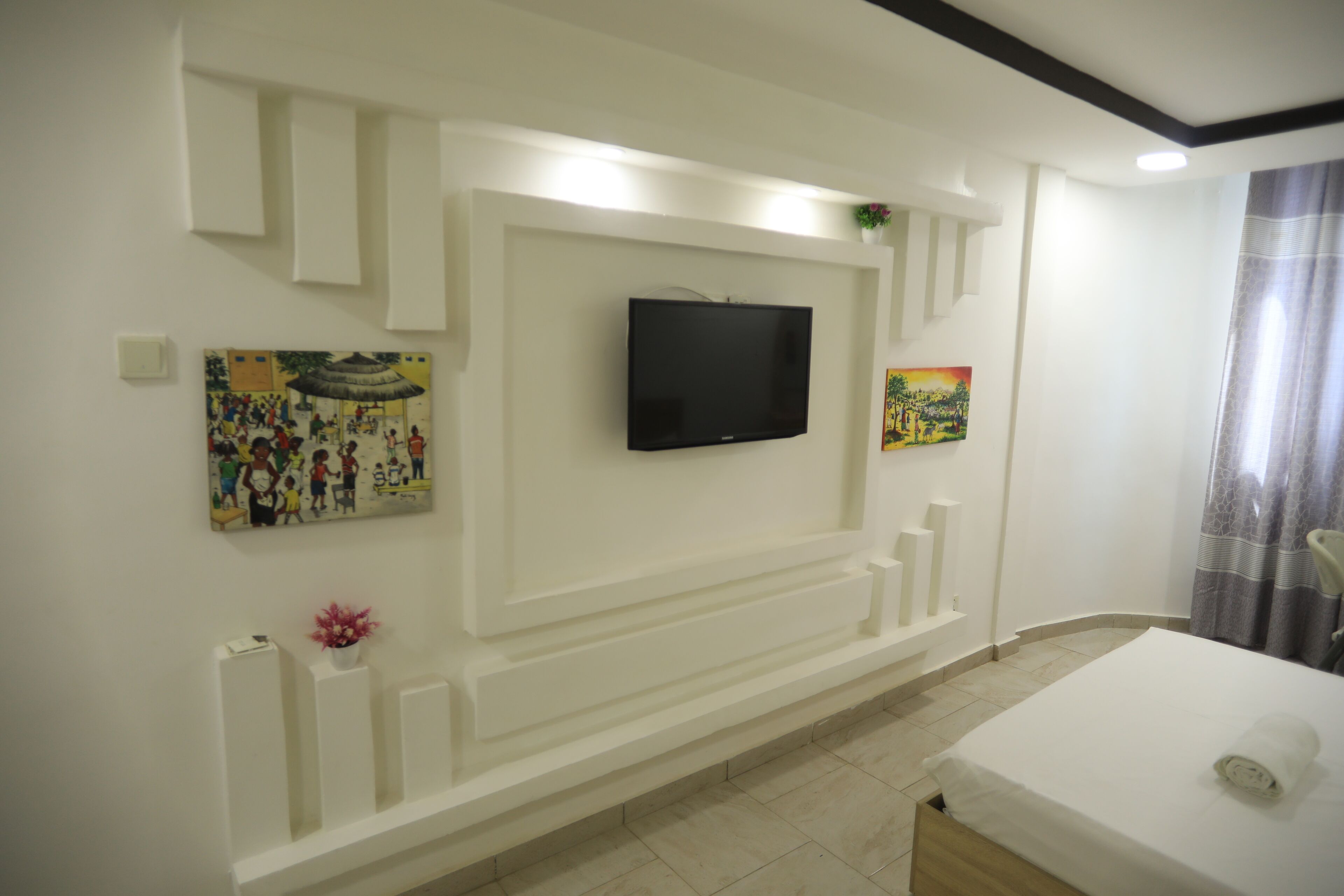 Habitación de hotel con televisión de pantalla plana en la pared, cama con colchón blanco y una toalla enrollada, y una pequeña planta en maceta sobre la mesita de noche.
