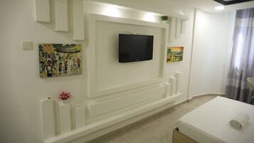 Habitación de hotel con televisión de pantalla plana en la pared, cama con colchón blanco y una toalla enrollada, y una pequeña planta en maceta sobre la mesita de noche.