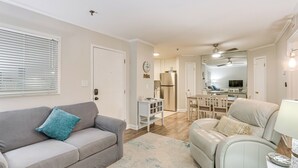 A living room with a grey sofa, a white armchair, and a coffee table. There is a kitchen in the background with a refrigerator and a dining table. - 'wright by the Beach': Hilton Head Condo w/ Perks (Hilton Head Island)