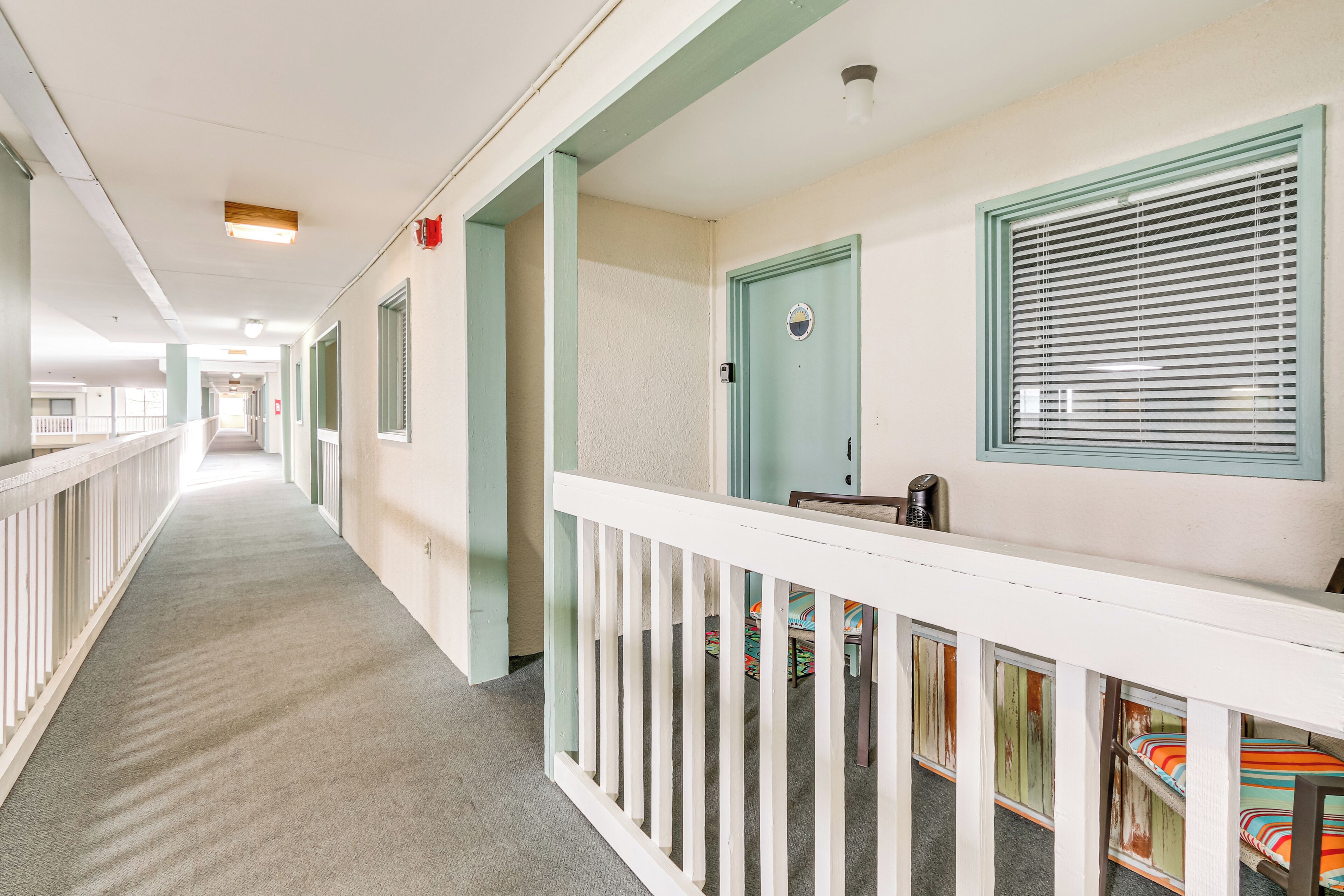 A corridor with a white railing, a door with a green frame, and a window with blinds.