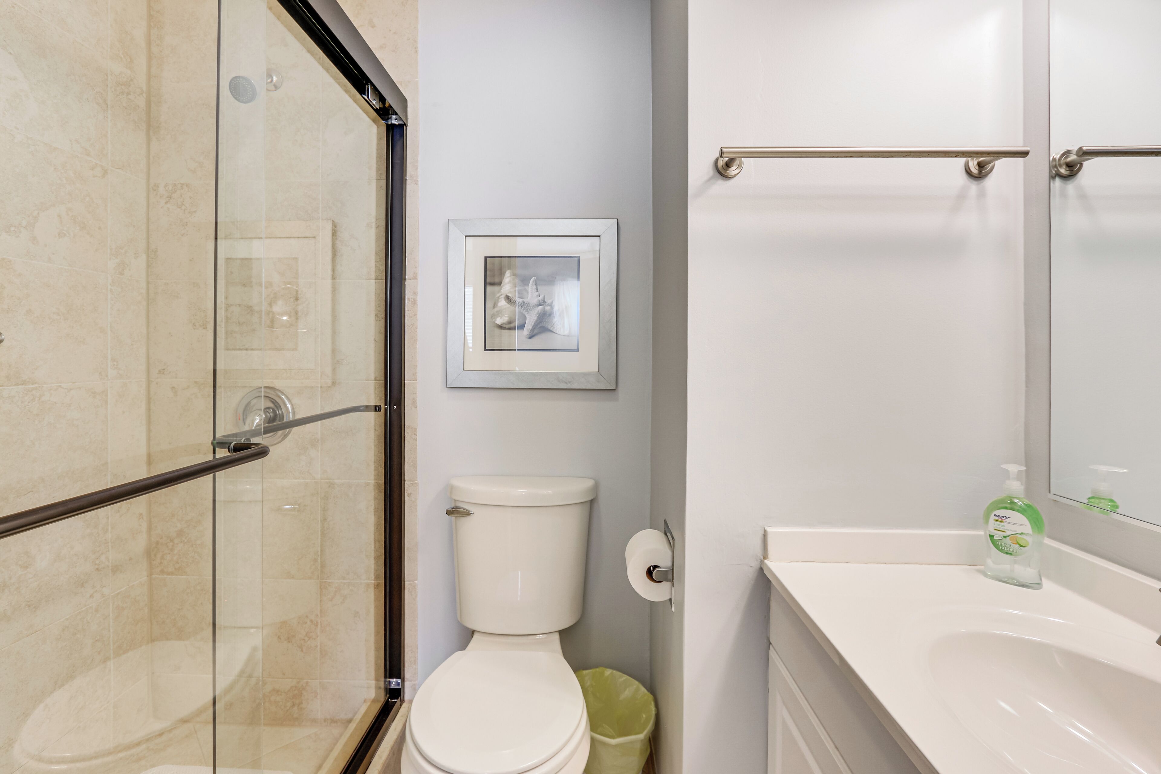 A bathroom with a glass-enclosed shower, a toilet, a framed picture on the wall, and a sink with a soap dispenser.