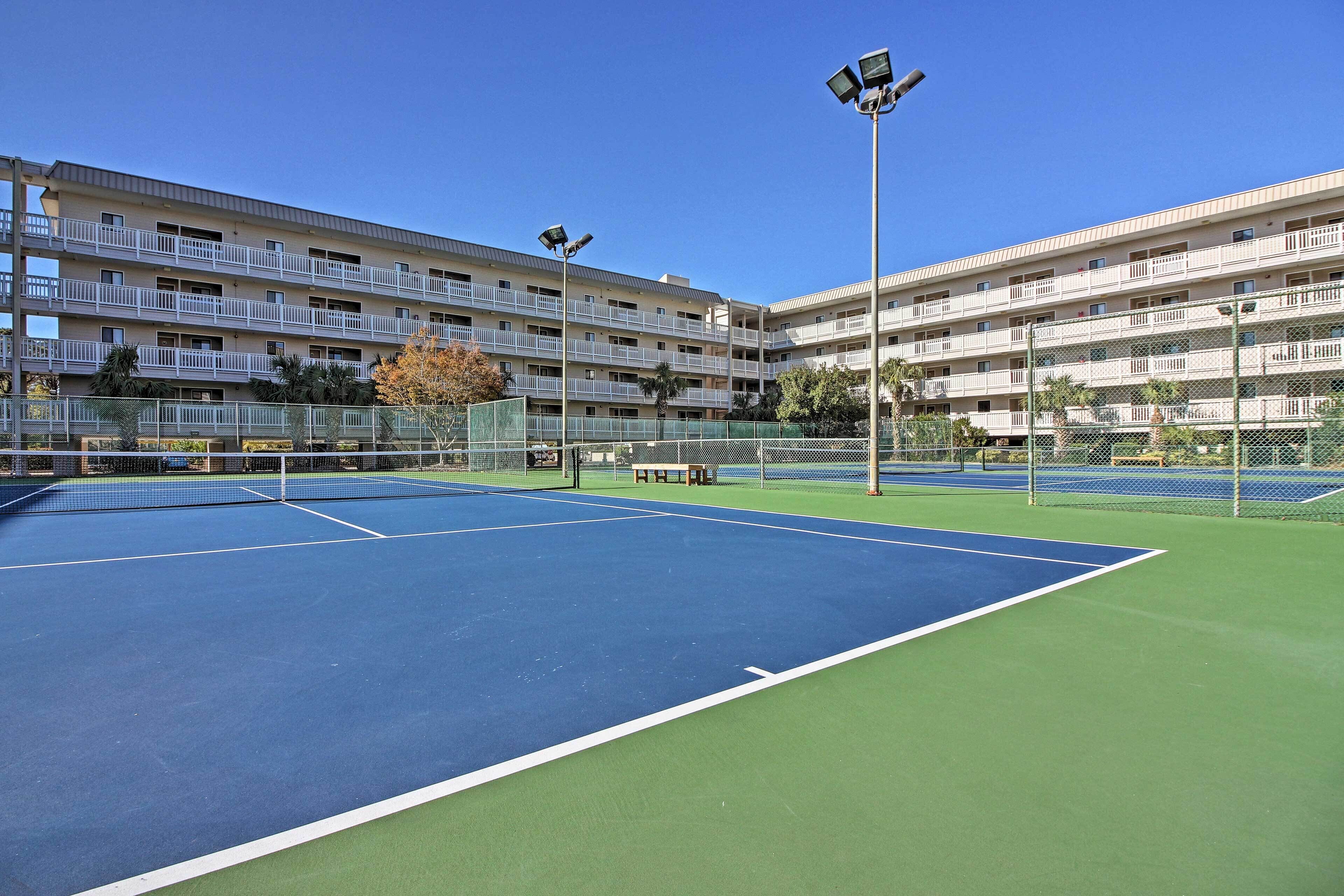 An outdoor tennis court with a net, surrounded by a fence, and adjacent to a multi-story building with balconies.