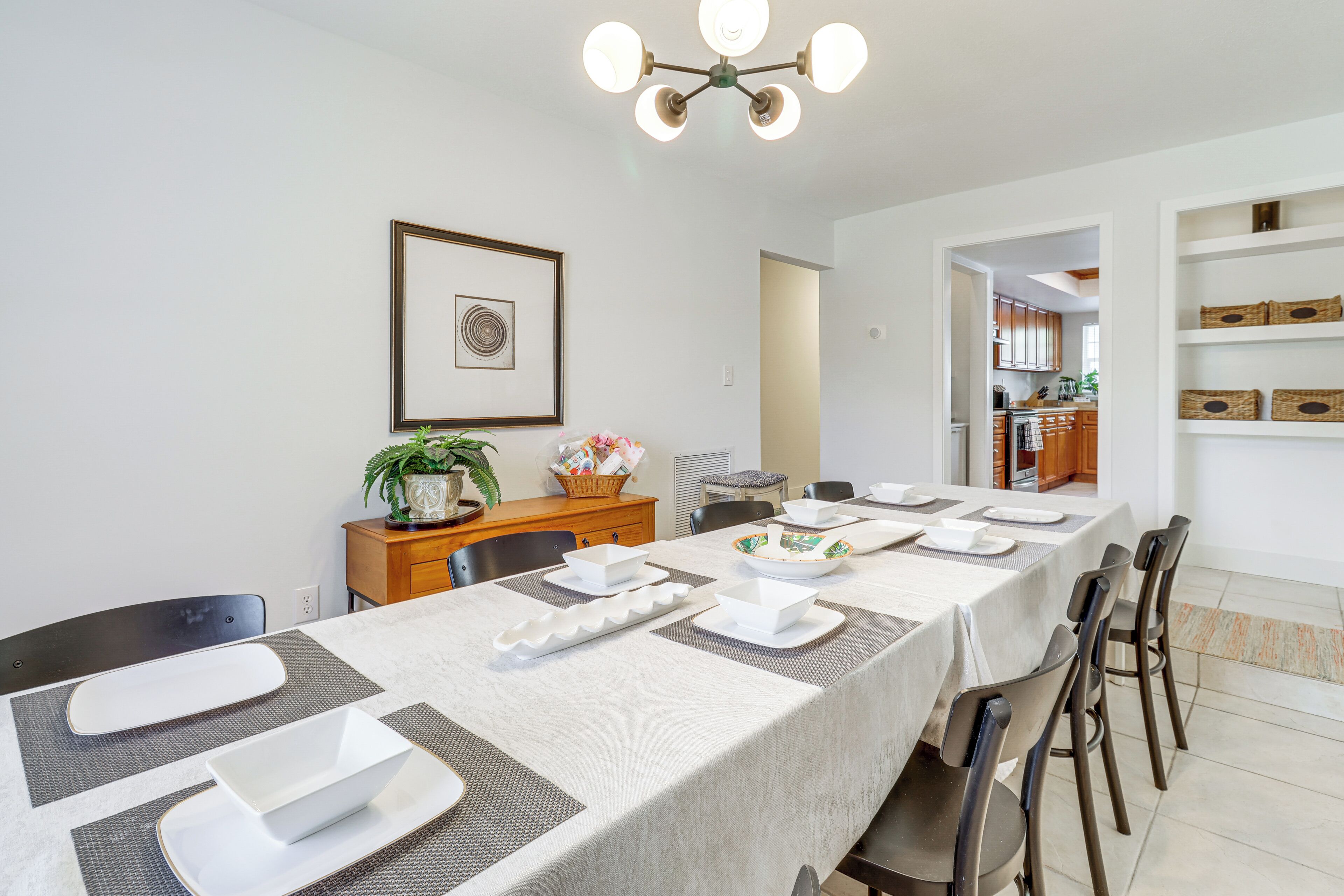 A dining area with a long table set for four, surrounded by chairs, with a kitchen visible in the background.