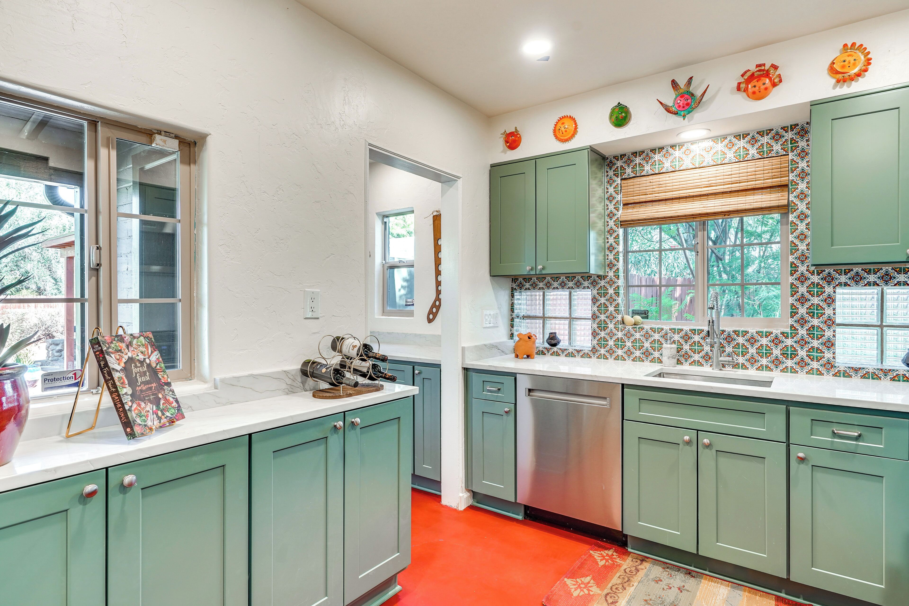 A kitchen with teal cabinets, a stainless steel dishwasher, and a window with a patterned tile backsplash.
