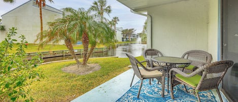 A covered patio with wicker furniture and a blue patterned rug, overlooking a body of water and palm trees.