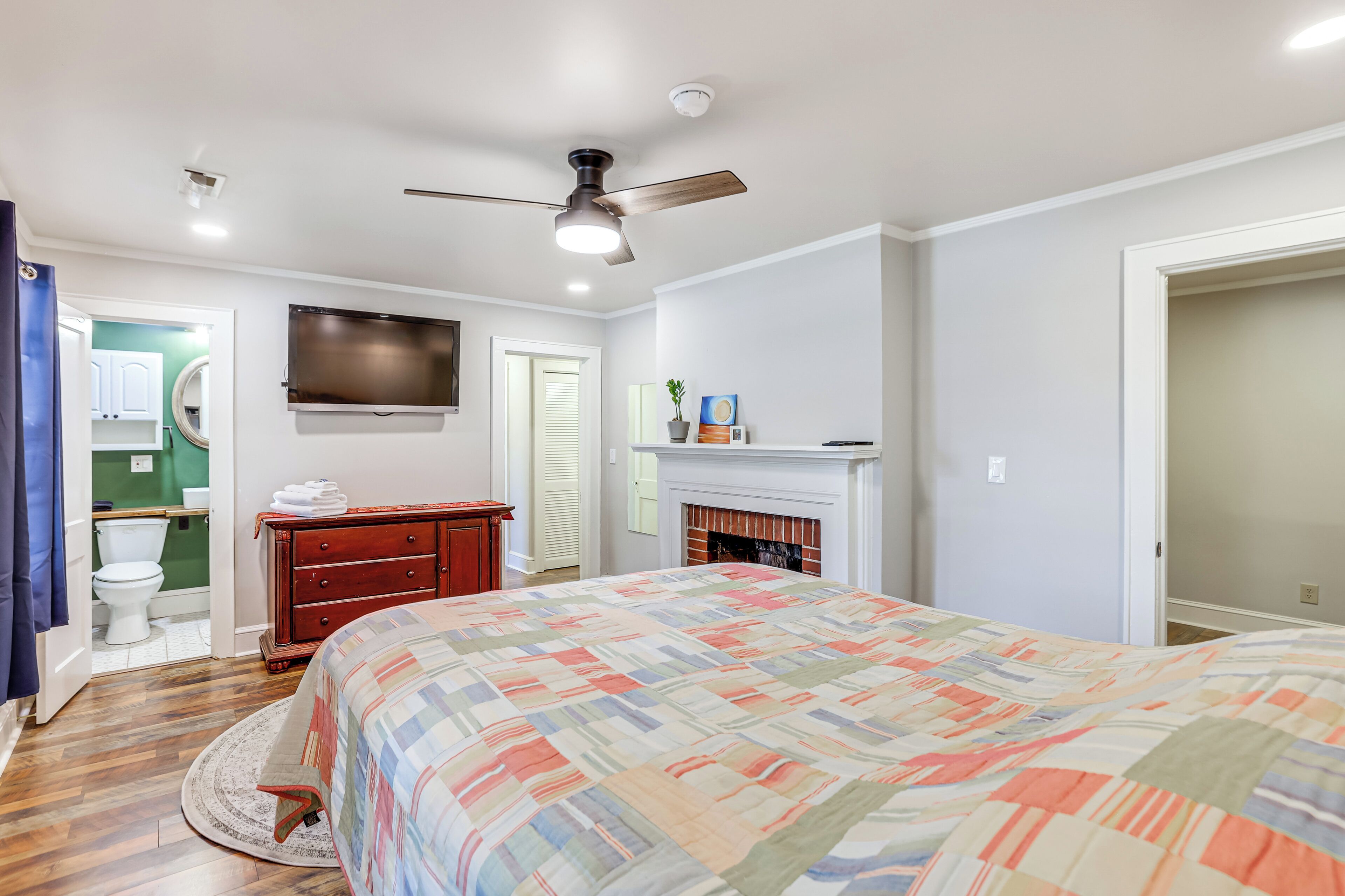 A bedroom with a bed, a ceiling fan, a fireplace, a TV, and a bathroom with a toilet and sink.