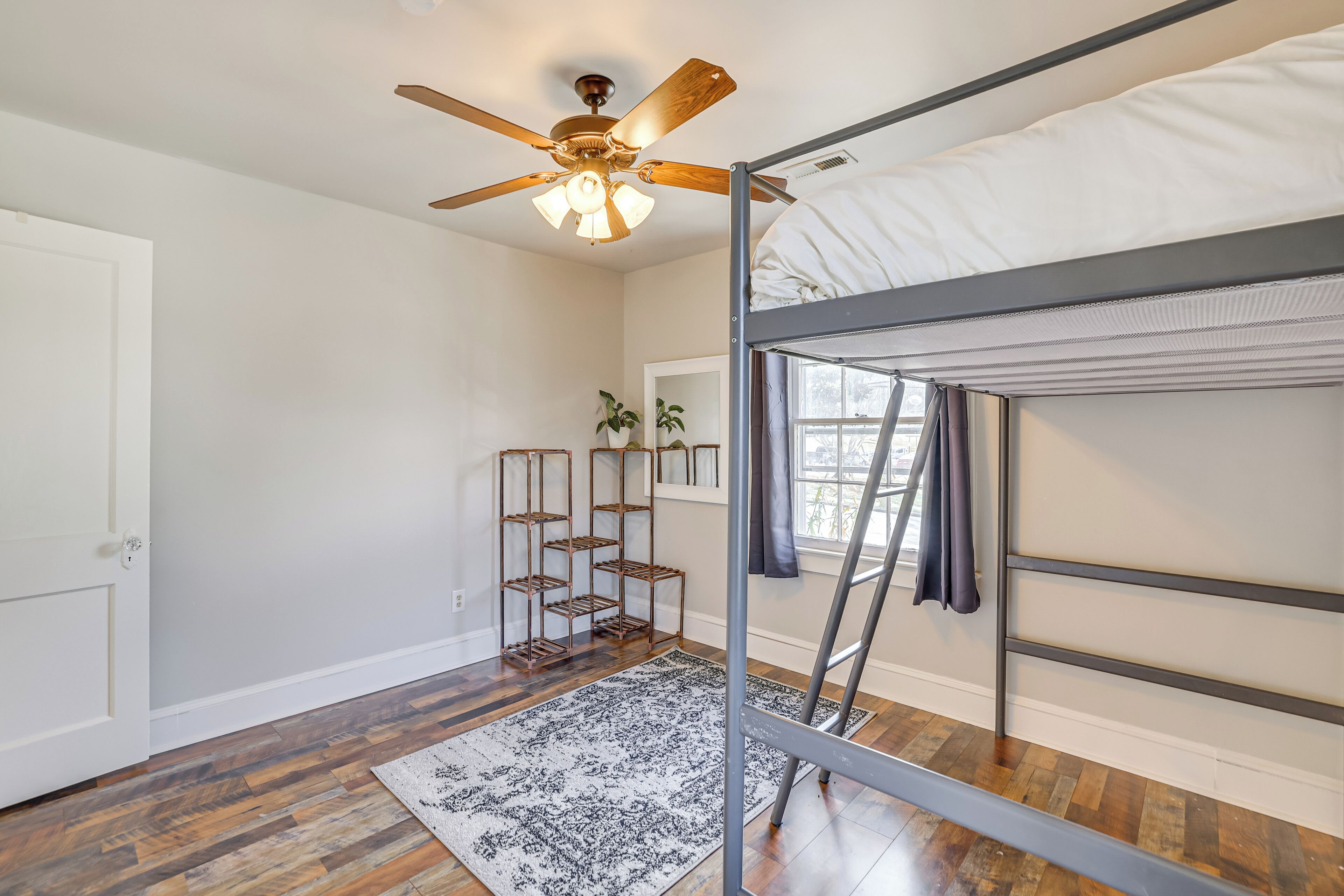 A room with a bunk bed, a ceiling fan, a window with curtains, a plant, and a patterned rug.