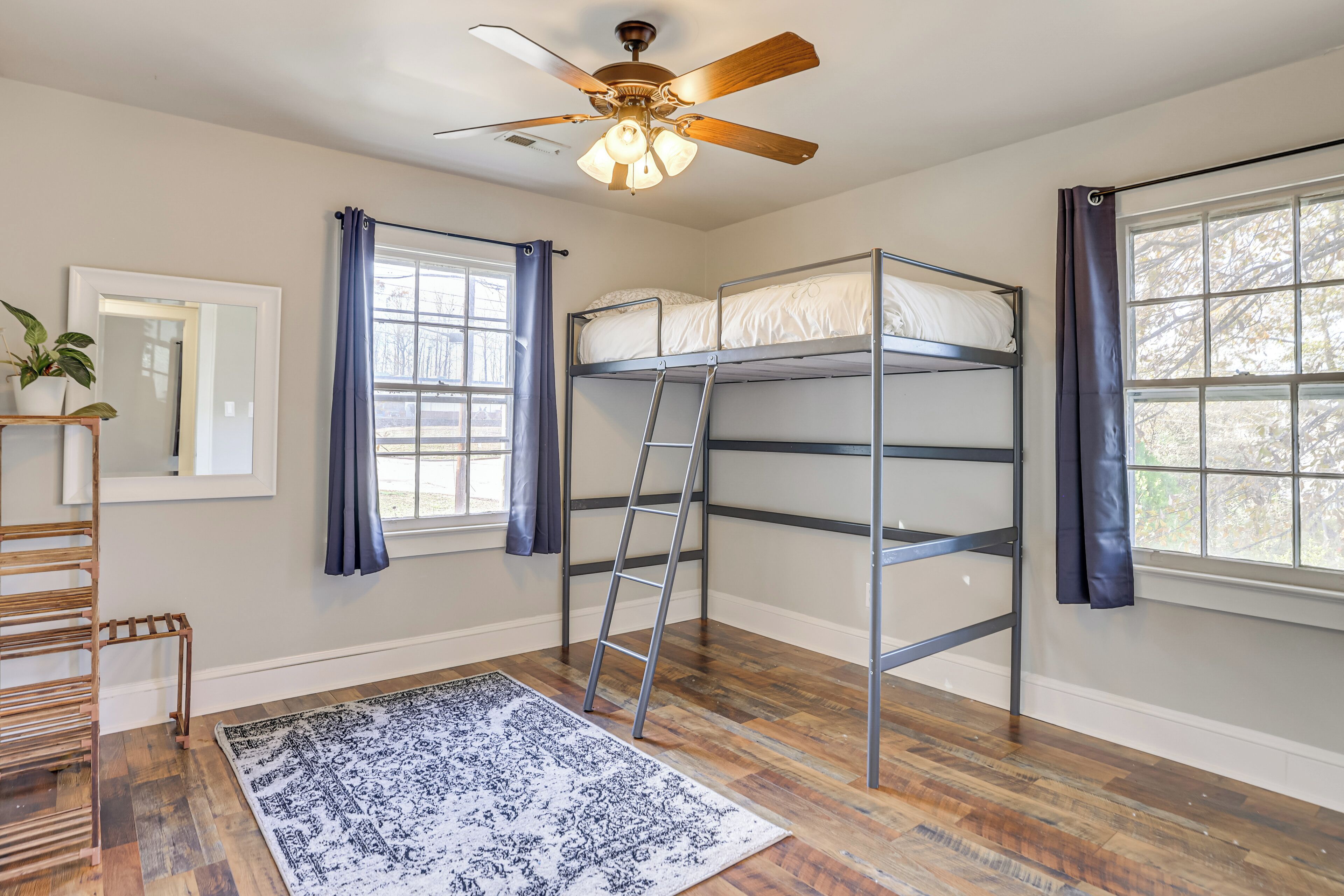Une chambre avec des lits superposés, un ventilateur de plafond, une fenêtre avec des rideaux, un plancher en bois et un petit tapis.